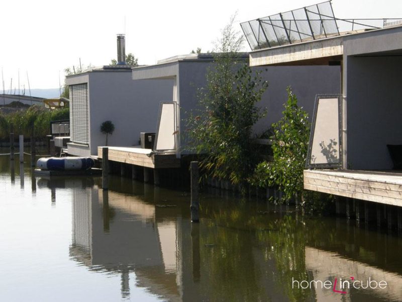 Detail vstupu, příjezdu a parkování Lake apartmánů, plechové fásádní obložení a krycí parkovácí stání s prefabrikovanou železobetonovou pergolovou konstrukcí a vetknutým dřevěným úložným prostorem.