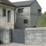 Plné betonové oplocení je v souladu s tmavým domem. Beton nádherně doplňují ocelová vrata ozdobená názvem ateliéru.