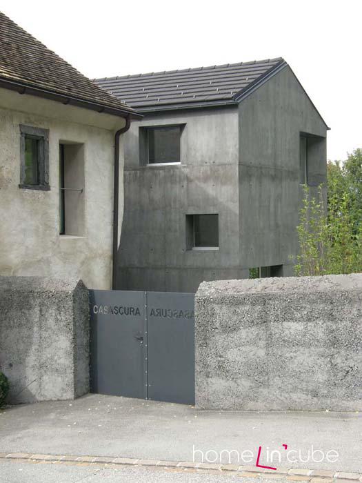 Plné betonové oplocení je v souladu s tmavým domem. Beton nádherně doplňují ocelová vrata ozdobená názvem ateliéru.